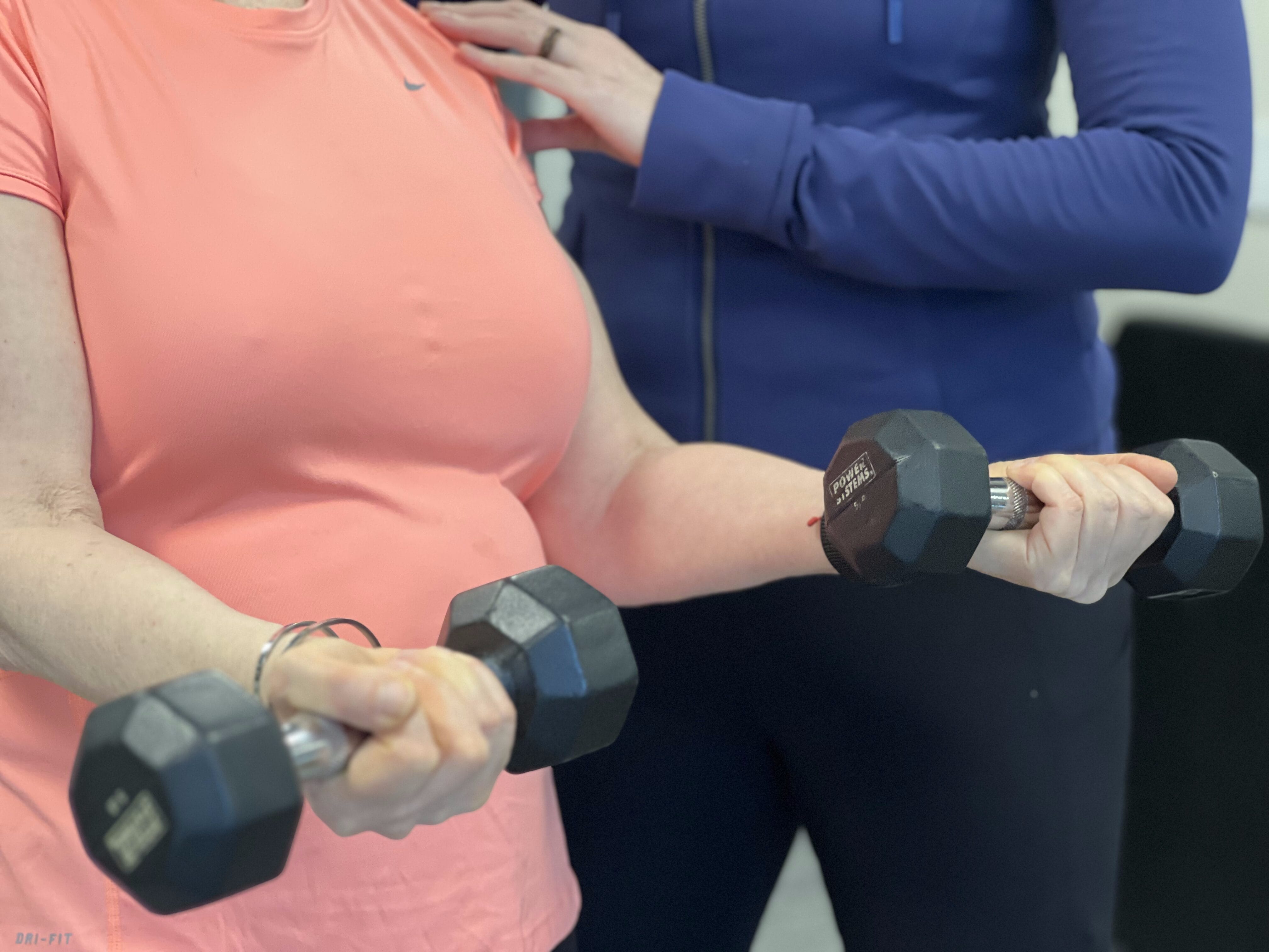 A person in a pink shirt performs bicep curls with dumbbells while another in a blue jacket provides support during functional strength training in Chicago.