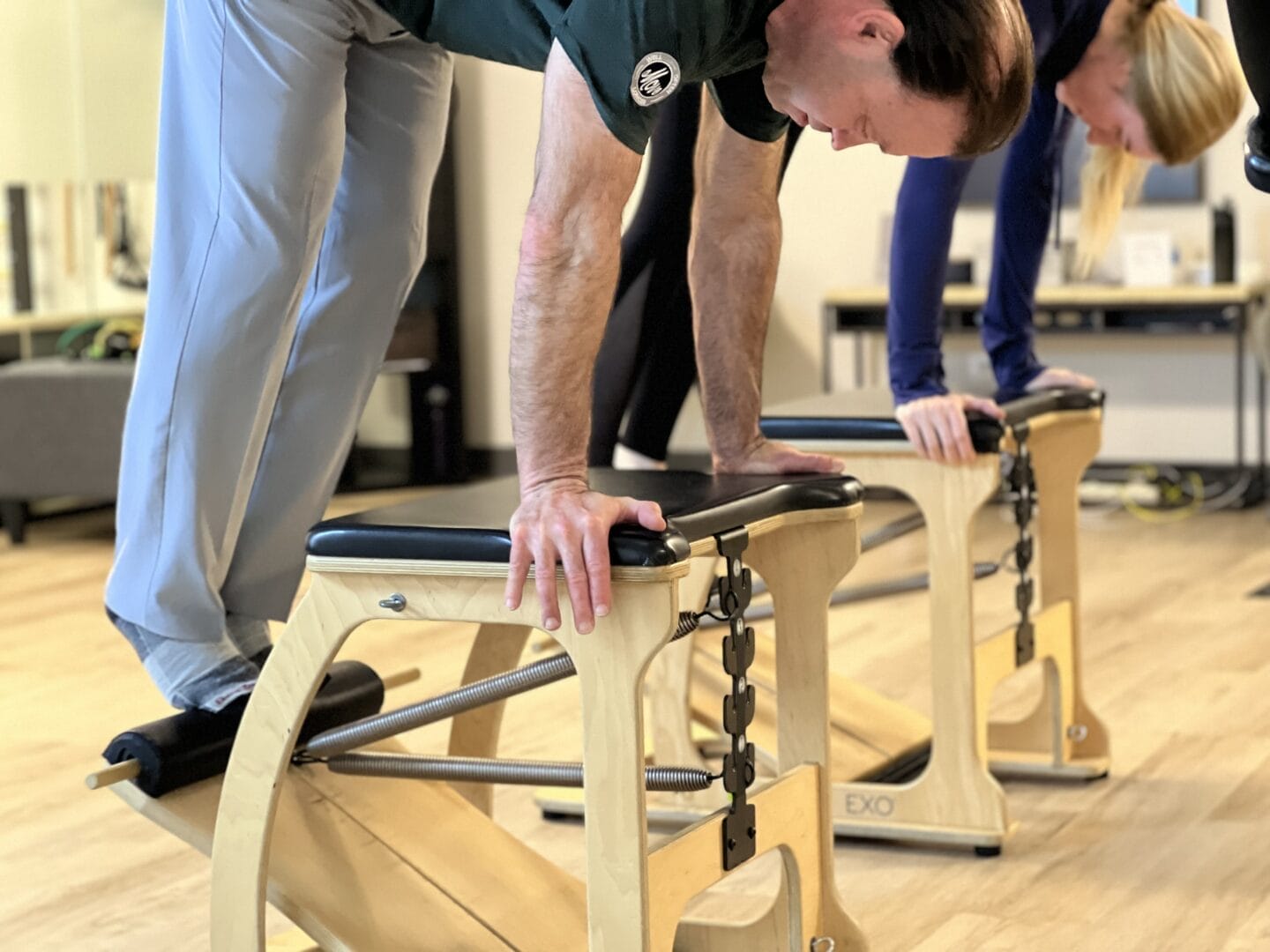Two people performing an exercise while leaning on wooden Pilates chairs in a fitness studio, enjoying Small Group Fitness Classes in Chicago.