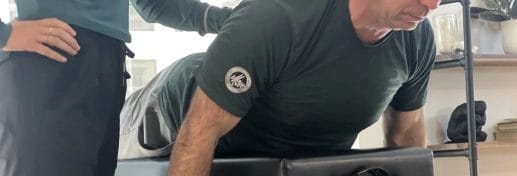 A man lies face down on a padded table while another person stands beside him, providing support during what appears to be a physical therapy or Pilates for Rehab in Chicago session.