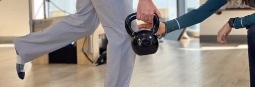 Person balances on one leg while holding a black kettlebell, assisted by another reaching toward them in a room with wooden floors—showcasing Functional Strength Training in Chicago.